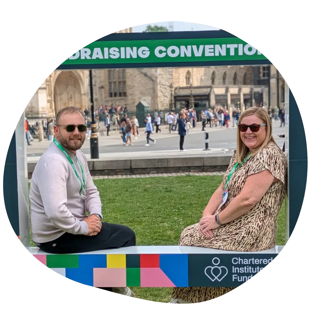 A smiling man and woman wearing sunglasses pose in a large frame outside the QEII Centre London